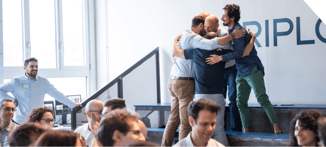 A group of men embraces in celebration on stairs, while an audience of seated individuals watches with smiles in a well-lit, modern indoor setting.